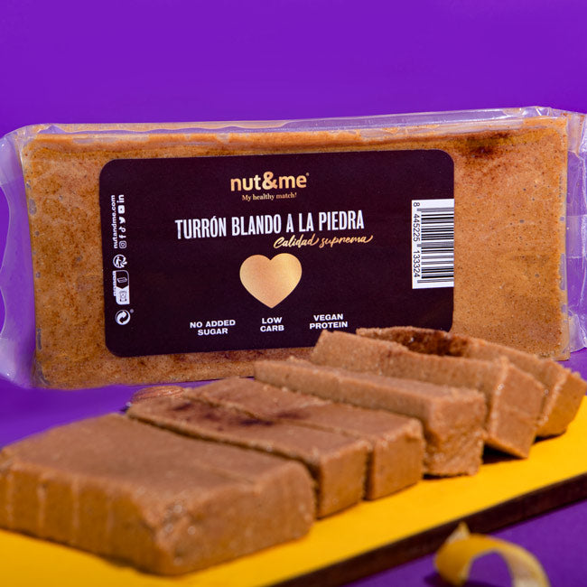 Una fotografía vibrante muestra un paquete de turrón "nut&me" con un fondo púrpura. El turrón, de tono dorado y de apariencia suave, está parcialmente sacado del paquete y cortado en rebanadas rectangulares que yacen sobre una superficie amarilla. En el paquete, se destaca el texto "Turrón Blando a la Piedra" y se resaltan etiquetas como "No Added Sugar", "Low Carb" y "Vegan Protein". Un corazón dorado en el centro del paquete simboliza "Calidad Suprema".